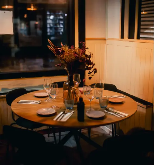 Mesa redonda de madera preparada para la cena en el comedor de Nakama, con copas, flores secas y luz cálida